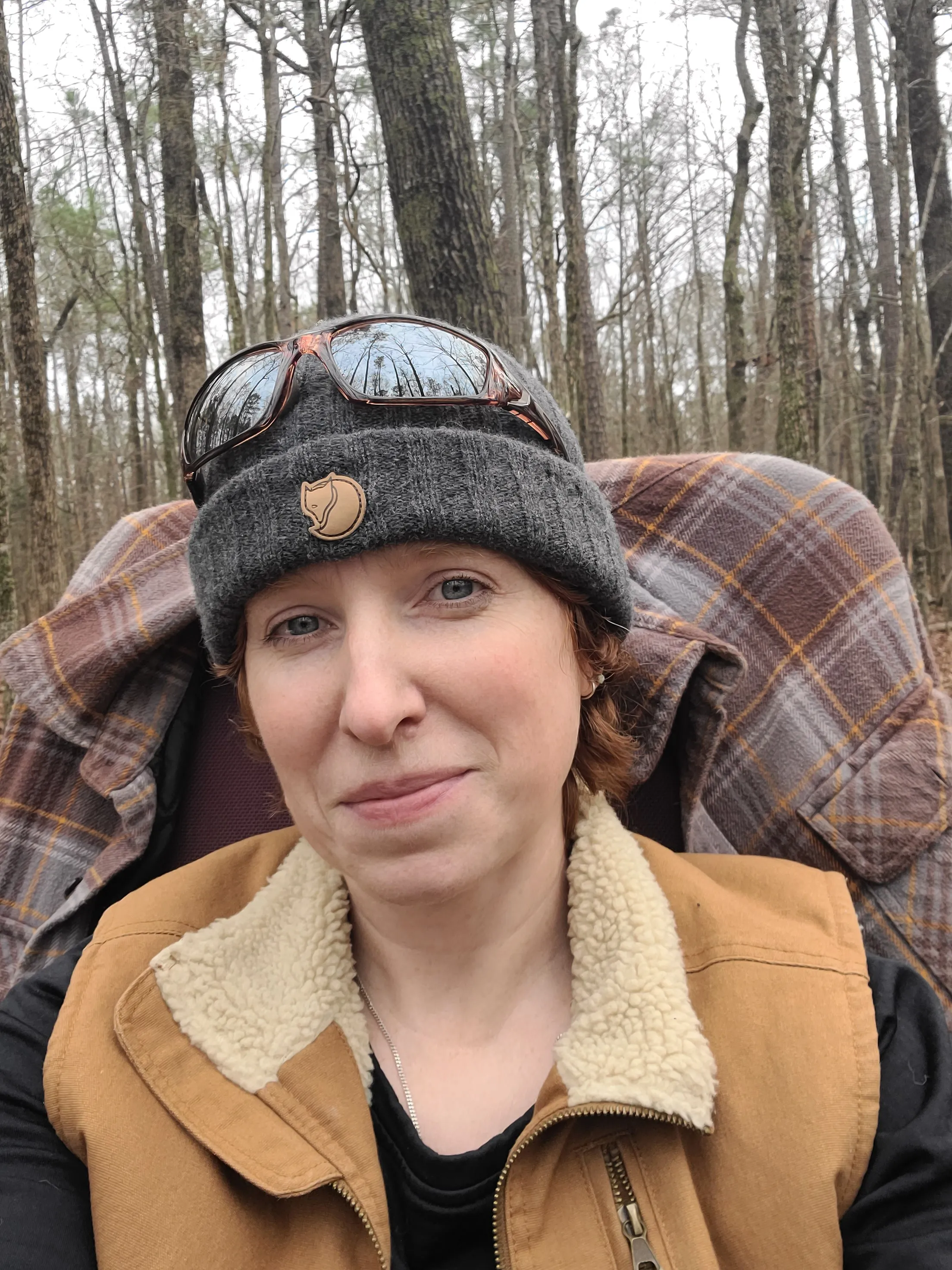 Georgia Leigh Carroll, in a beanie and plaid, sitting in winter woods.